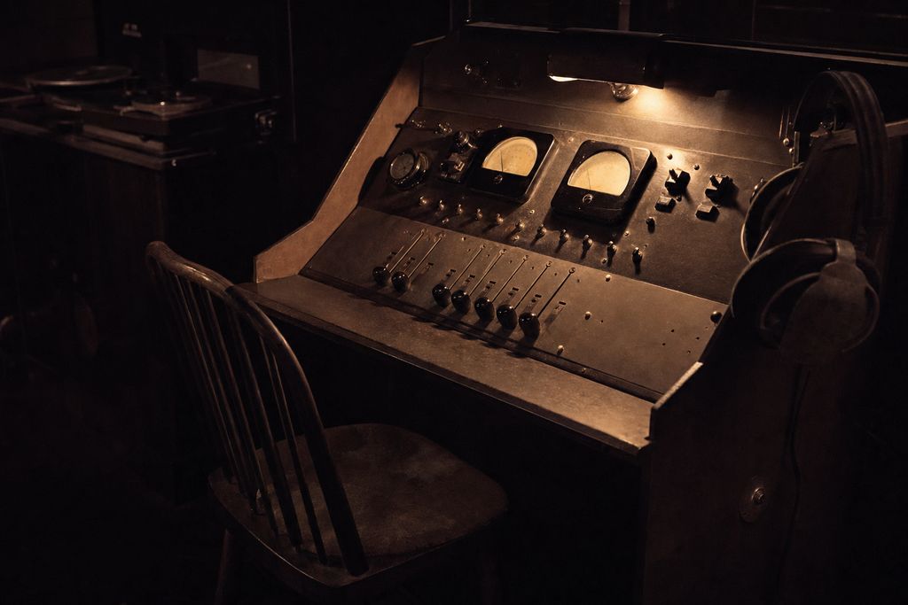 A radio continuity desk with rotary faders and two large VU meters, headphones hooked over the corner and an empty chair pushed back