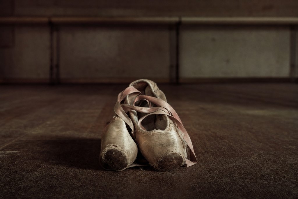 A pair of worn ballet shoes on the sprung floor of a rehearsal room, a barre out of focus behind.